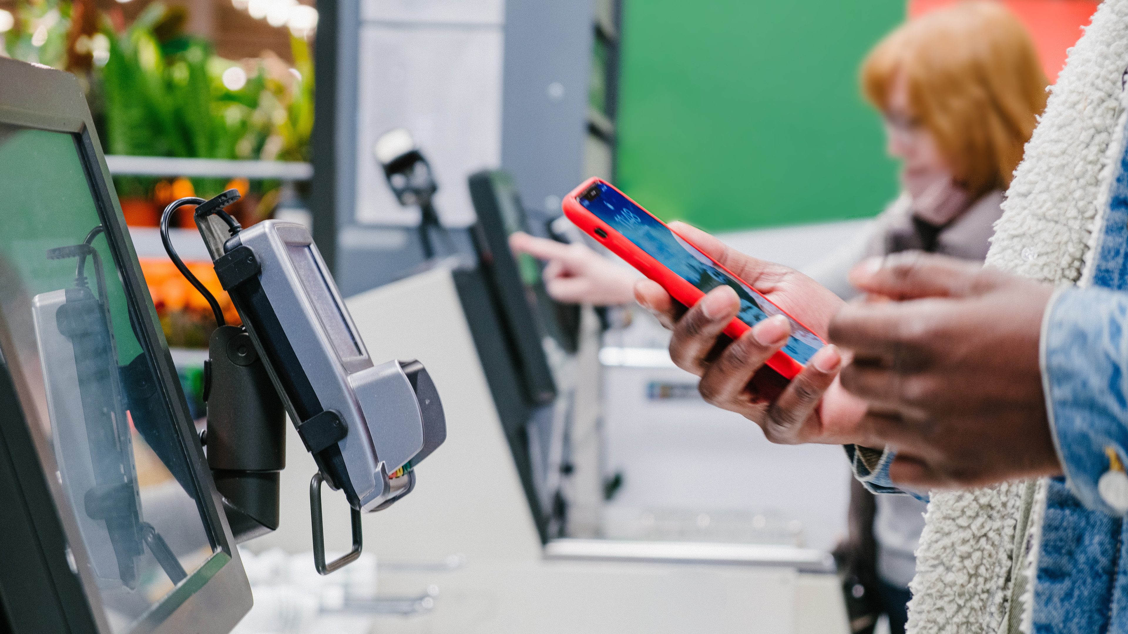 Black male person in warm denim jacket uses smartphone to pay for purchase at self-checkout point in supermarket close view