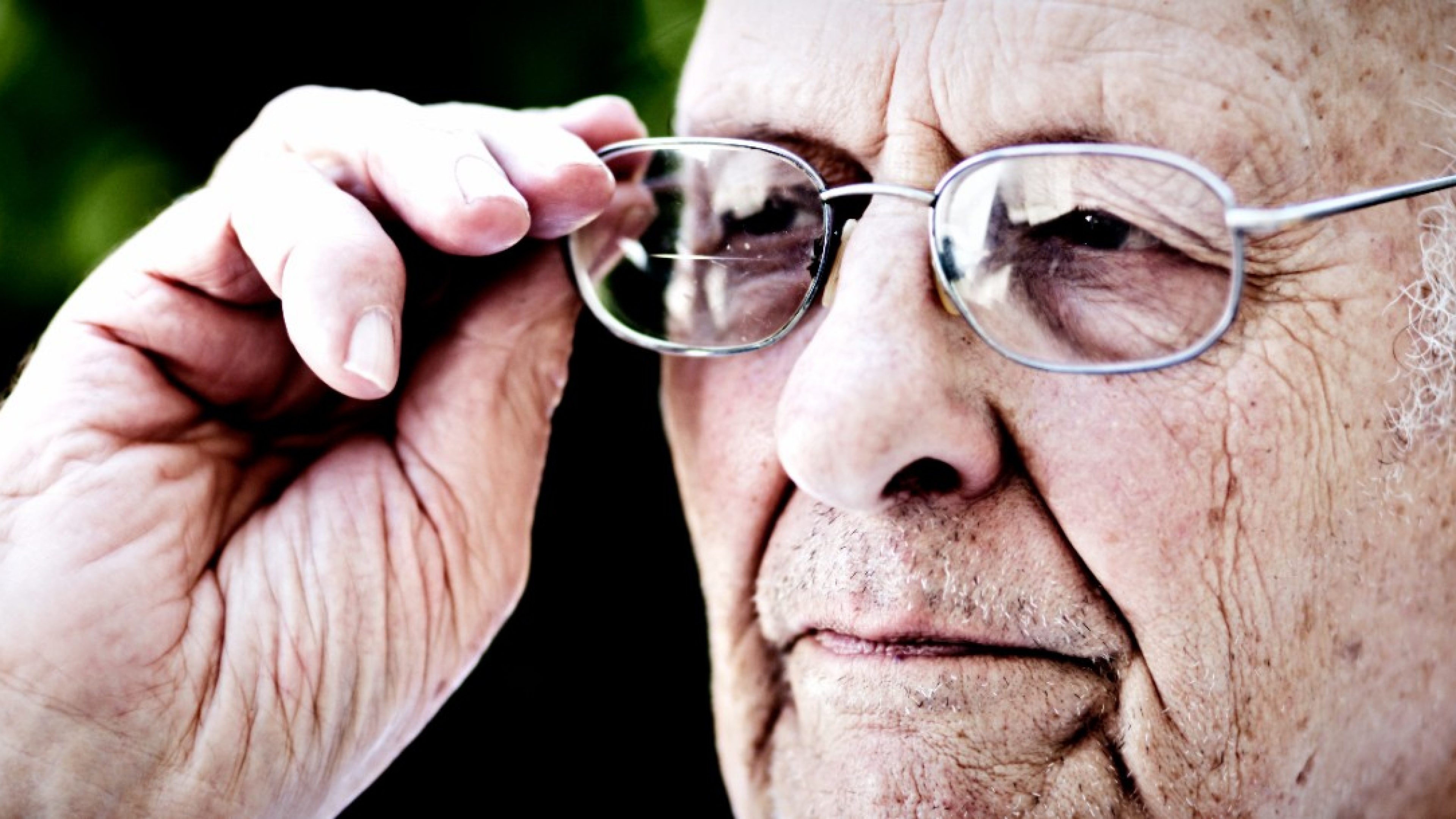 This 90 year old man adjusts his eyeglasses as he stands in his garden, looking serious.