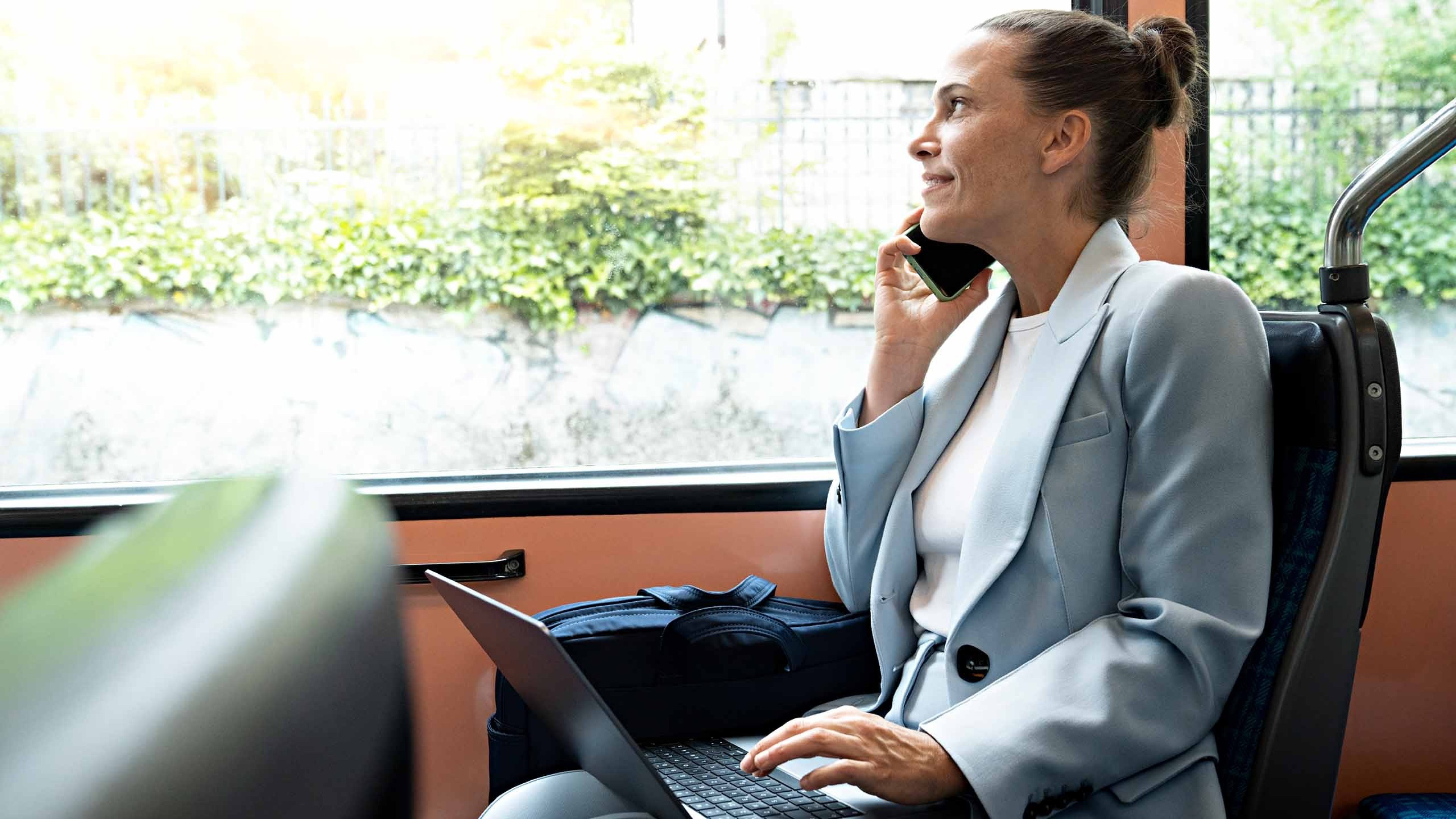 Eine Frau sitzt mit dem Laptop auf den Knien im Tram und schaut telefonierend aus dem Fenster.