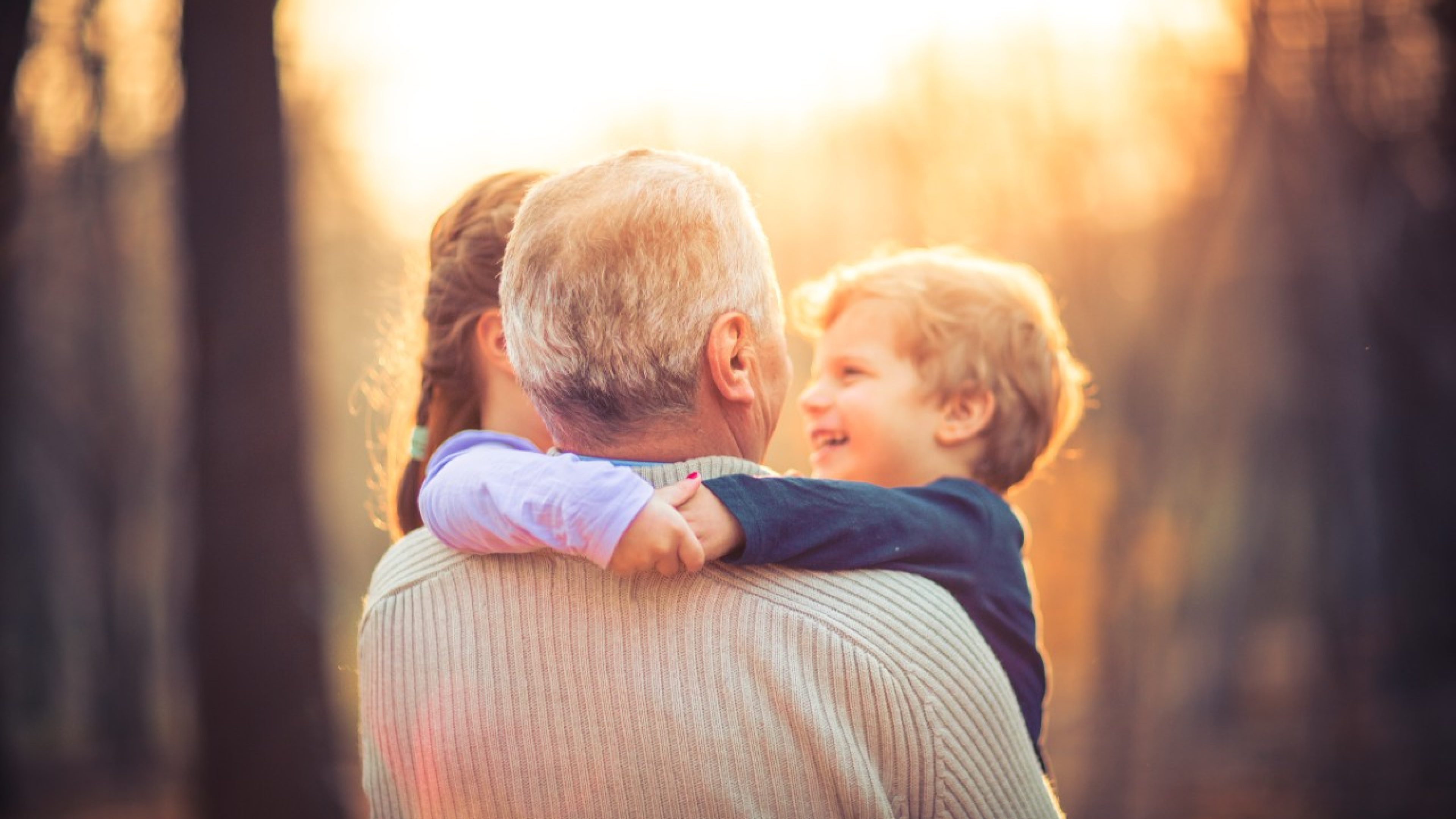 Picture of a grandfather playing with his two grand children a five year old granddaughter and three year old grandson in the park. The old man is wearing a khaki sweater and a blue shirt with long sleeves. Boy and girl wrapped their hands around grandfather. The little girl is wearing a pink blouse and pink sweatpants and boy is wearing blue blouse. They are smiling and having fun. The lens flare is behind them.